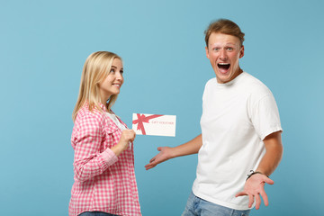 Young overjoyed couple two friends guy girl in white pink empty blank t-shirts posing isolated on pastel blue background in studio. People lifestyle concept. Mock up copy space. Hold gift certificate.