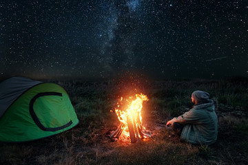 Camping man sits by the fire at night against the background of the starry sky. The concept of travel, tourism, camping.