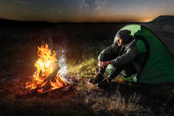 Camping man sits by the fire at night against the background of the starry sky. The concept of travel, tourism, camping.