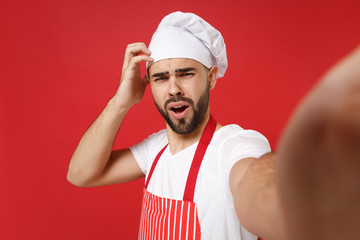 Close up of preoccupied bearded chef cook baker man in striped apron toque chefs hat posing isolated on red background. Cooking food concept. Mock up copy space. Doing selfie shot on mobile phone.