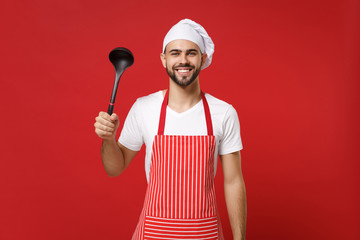 Cheerful young bearded male chef cook or baker man in striped apron toque chefs hat posing isolated on red wall background. Cooking food concept. Mock up copy space. Holding soup black ladle dipper.