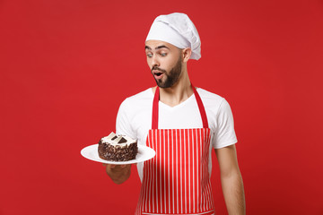 Shocked young bearded male chef cook or baker man in striped apron white t-shirt toque chefs hat posing isolated on red background. Cooking food concept. Mock up copy space. Holding plate with cake.