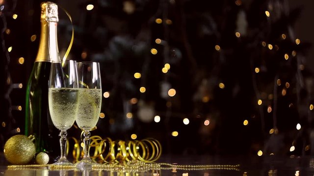 Two glasses and green bottle with champagne, golden decor, balls, serpentine are on table. Festive decorative garland, warm yellow light bulbs are blinking on background. New year, christmas mood. 