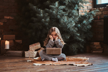 Grumpy unhappy girl is sitting next to christmas tree with big gift.