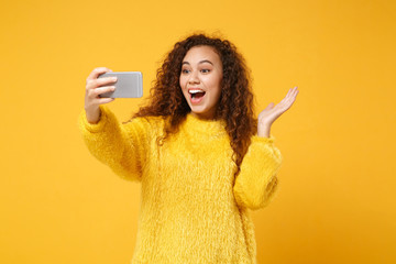 Excited young african american girl in fur sweater posing isolated on yellow orange wall background. People lifestyle concept. Mock up copy space. Doing selfie shot on mobile phone, spreading hands.