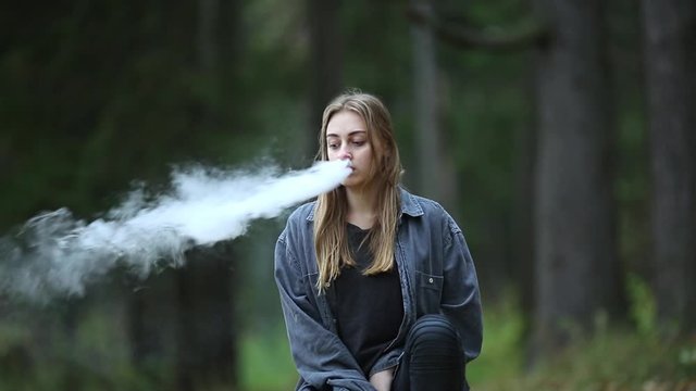 Vape teenager. Young beautiful white teenage girl in casual clothes vaping electronic cigarette on the street in the park in the autumn evening. Bad habit. Vaping activity.