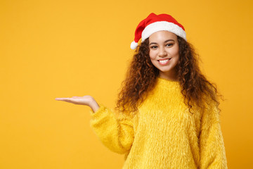 Smiling young african american Santa girl in fur sweater, Christmas hat isolated on yellow wall background. Happy New Year 2020 celebration holiday concept. Mock up copy space. Pointing hand aside.