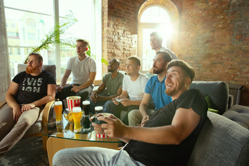 Group of excited friends playing video games at home. Caucasian male gamers or fans spending time and having fun together at home. Emotional watching gameplay. Modern technologies, friendship, weekend