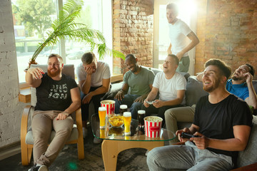 Group of excited friends playing video games at home. Caucasian male gamers or fans spending time and having fun together at home. Emotional watching gameplay. Modern technologies, friendship, weekend