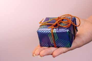 Wrapped gift box with a bow in the hand of a girl with long nails. Blue and green wrapping paper, orange lace, pink background. Copy space.