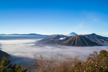 Mount Bromo volcano, island of East Java, Indonesia. Clouds cover the valley floor; Luhur Poten Temple at the foot of the cone. Gas from another volcano in the background rising against the blue sky.