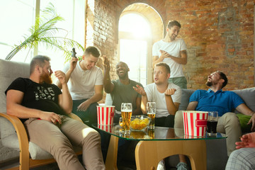 Group of excited friends playing video games at home. Caucasian male gamers or fans spending time and having fun together at home. Emotional watching gameplay. Modern technologies, friendship, weekend