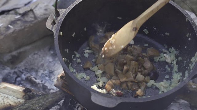 Frying onion and girolle (Cantharellus cibarius), Porcini (Boletus) mushroom for risotto in cast iron pot on an open campfire