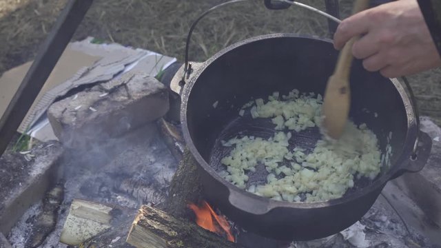 Frying onion for girolle (Cantharellus cibarius), Porcini (Boletus) mushroom and chicken risotto in cast iron pot on an open campfire
