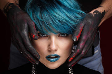 Ears of beautiful girl with short blue fashionable hairstyle and blue lipstick covered by men's hands in black, pink and red paint on a dark background. Hear no evil concept. Close-up studio photo