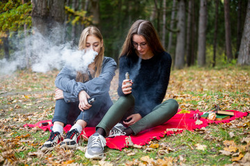 Vape lgbt teenagers. Bisexual lesbian young caucasian teenage girls in casual clothes vape electronic cigarette near the river on the street in the park in the autumn evening. Bad habit.
