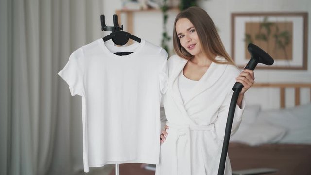 Beautiful young woman shows steamer for clothes and looks at camera