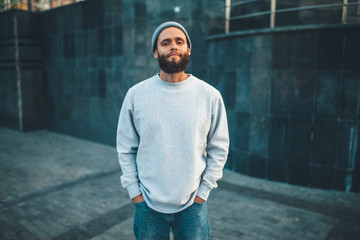 City portrait of handsome hipster guy with beard wearing gray blank hoodie or hoody and hat with...