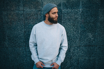 City portrait of handsome hipster guy with beard wearing gray blank hoodie or hoody and hat with...