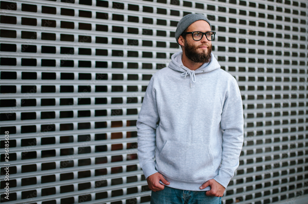 Wall mural City portrait of handsome hipster guy with beard wearing gray blank hoodie or sweatshirt and hat with space for your logo or design. Mockup for print - Wall murals