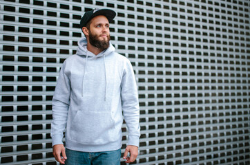 City portrait of handsome hipster guy with beard wearing gray blank hoodie or sweatshirt and hat with space for your logo or design. Mockup for print