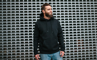 City portrait of handsome hipster guy with beard wearing black blank hoodie or sweatshirt and hat...