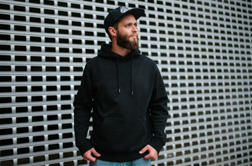City portrait of handsome hipster guy with beard wearing black blank hoodie or sweatshirt and hat...