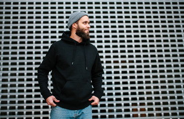 City portrait of handsome hipster guy with beard wearing black blank hoodie or sweatshirt and hat...