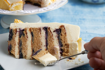 Close-up of a piece of layer cake with cream cheese, lemon and blueberries. You can see a hand holding a fork that takes some cake.