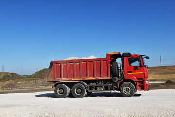 A large red truck loaded with sand. A truck carries building materials on a highway rack. Roadworks are going everywhere, new asphalt is laid.