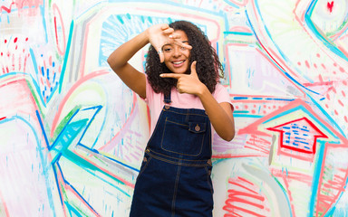 young pretty african american woman feeling happy, friendly and positive, smiling and making a portrait or photo frame with hands against graffiti wall