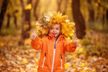 Portrait of little toddler boy in crown of yellow leaves standing in colorful autumn park or forest and showing two thumbs up. Seasonal games outdoors, fun and happy carefree childhood concept