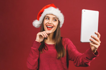 Ney Year sale! Attractive woman in santa hat and sweater holding digital tablet isolated on red.