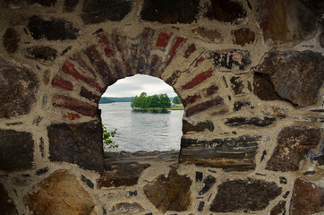 The fortress wall is made of natural stone with a loophole. Stones of different colors and shapes. Behind the wall you can see a lake and a shore with trees. Background.