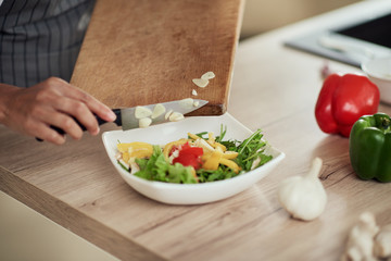 Close up mixed race woman in apron holding cutting board and putting chopped garlic in bowl with vegetables.