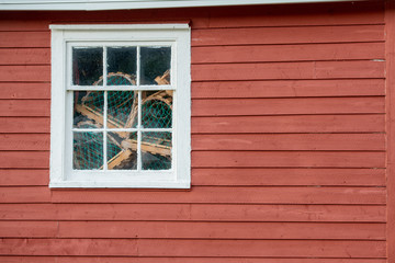 A red exterior clapboard wall with a small nine pane window with white trim. There are lobster traps in the window made of wood and green rope and netting. The old wall is textured and worn.