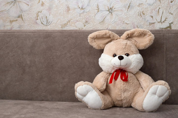 Plush rabbit sitting on a brown sofa. Beige toy rabbit with a red bow. Place for text. Romantic card