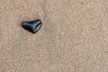 Fototapeta na wymiar A black stone or beach rock on sits on a beach filled with small grains of sand. There's a pink tint to the sand. The stone is wet and in the shape of a heart.