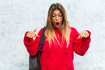 young pretty student feeling shocked, open-mouthed and amazed, looking and pointing downwards in...