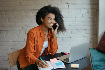 Young businesswoman using laptop, talking on mobile phone, taking notes, working startup project in...