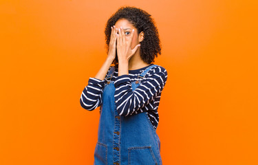 young pretty black woman covering face with hands, peeking between fingers with surprised...