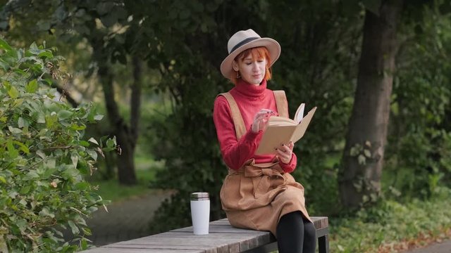 Young attractive redhead woman in a hat and sweater reading a book and drinking coffee from a thermocup on a bench in a natural park. Autumn cool day, green and yellow trees.