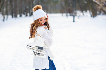 Beautiful woman weared in white sweater and hat with ice skates on the back walks in winter snowy park.