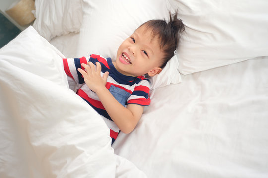 Cute little happy smiling Asian 3 - 4 years toddler boy child waking up in bed, Cheerful kid lying on bed looking at camera on lazy day, Morning awakening, Good morning at home concept