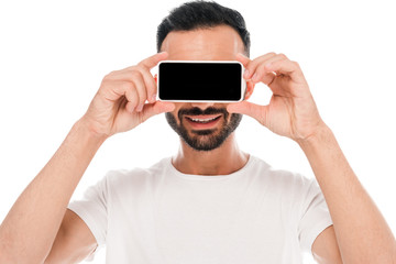 bearded man covering eyes while holding smartphone with blank screen isolated on white