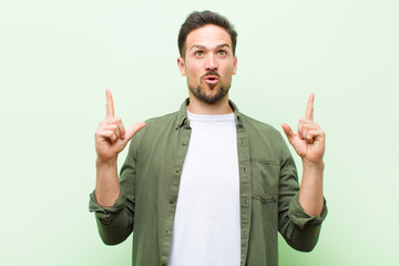 young handsome man looking shocked, amazed and open mouthed, pointing upwards with both hands to...