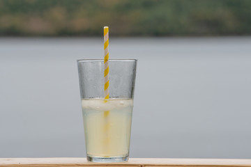 A tall glass of homemade lemonade with a yellow and white stripped paper straw in the glass. The glass is sitting on a table with the ocean behind it.