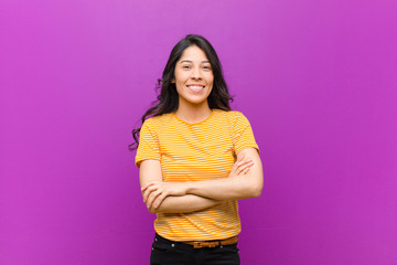 young pretty latin woman looking like a happy, proud and satisfied achiever smiling with arms crossed against purple wall