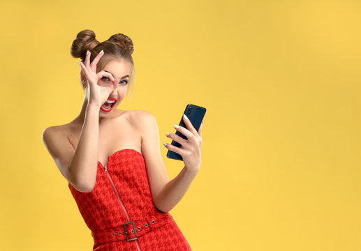 Happy girl holding a smartphone and showing okay sign wearing red dress on yellow background