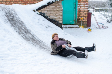 Mature senior woman riding sled from children slide on icey trek. Excited adult female haveing fun in winter like child. Traditional russian winter entertainment.